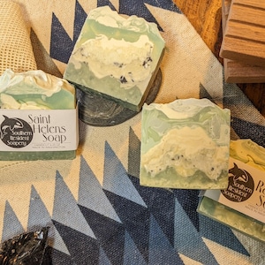 May include: Three bars of handmade soap with green and white swirls. The soap bars are labeled "Saint Helens Soap" and "Rainier Soap" with a logo of a whale. The soap bars are on a patterned fabric background.
