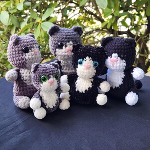 May include: A collection of five handmade crochet cat plushies in various colors. The cats have white paws, pink noses, and green or blue eyes. The color palette includes shades of gray, purple, and black. The plushies are arranged on a dark blue surface.