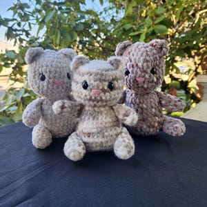 May include: Three handmade crochet teddy bears in shades of gray and brown. Each bear has black eyes and a pink nose. The bears are sitting on a dark blue surface, with green foliage in the background.
