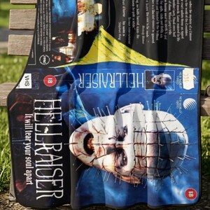 Könnte beinhalten: Eine rechteckige Fleecedecke mit dem Design des Hellraiser-Filmplakats. Der Druck zeigt die ikonische Figur Pinhead mit Nadeln im Kopf, den Titel Hellraiser, den Slogan It will tear your soul apart sowie weiteren Text zum Film.