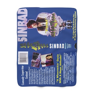 Peut inclure: Une couverture bleue avec le titre du film "Sinbad" et le mot "Shazam" en jaune et vert. L'image comprend une photo de Sinbad et le texte "A Wisecrackin' Genie With A Mean Attitude!"