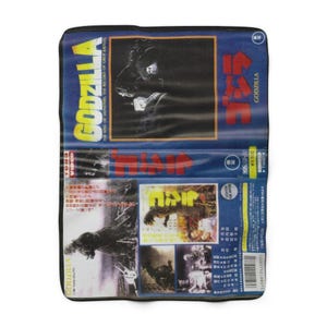 May include: A rectangular, blue and yellow Godzilla VHS tape. The tape features the Godzilla logo and Japanese text. The cover shows images of Godzilla and other monsters. The tape is in a black protective case.