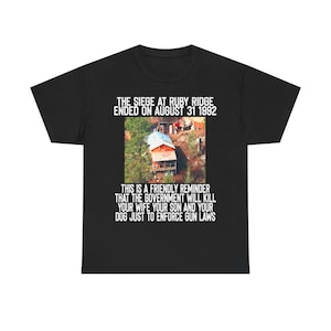 May include: Black t-shirt featuring an image of a building and text. The text reads "THE SIEGE AT RUBY RIDGE ENDED ON AUGUST 31 1992" and "THIS IS A FRIENDLY REMINDER THAT THE GOVERNMENT WILL KILL YOUR WIFE YOUR SON AND YOUR DOG JUST TO ENFORCE GUN LAWS."