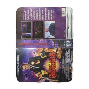 May include: A rectangular throw blanket featuring a VHS tape design with the title "Threat Level Midnight." The blanket has a purple and black color scheme with movie details and text, including "He Shoots... He Scores."