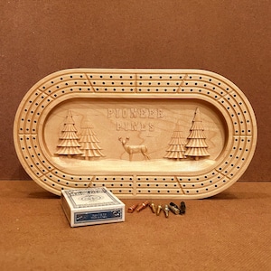 May include: A wooden cribbage board with a carved design of pine trees and a deer. The board is oval shaped and has a dark brown stain. The text "Pioneer Pines" is carved into the board. The board is accompanied by a deck of playing cards and a set of pegs.