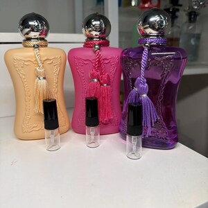 May include: Three perfume bottles in peach, hot pink, and translucent purple, each featuring a silver cap, a decorative tassel, and an ornate embossed frame design. Each bottle is paired with a small, clear glass sample vial with a black cap.