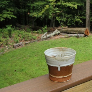 May include: A brown and white ceramic mug with a drip glaze sits on a wooden deck railing. The mug is in focus, while the background is a blurry green lawn and trees.
