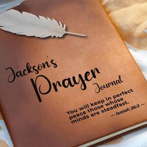 May include: A brown leather-bound journal with the words "Jackson's Prayer Journal" embossed on the front cover. The journal also features a quote from Isaiah 26:3, "You will keep in perfect peace those whose minds are steadfast."