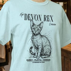以下が含まれることがあります： 水色の半袖Tシャツで、デボンレックス猫の黒い線画イラストが描かれています。テキストには「The Devon Rex Collective」「Quirky - Playful - Curious」「La Maison Du Chat」と記されています。Tシャツはマネキンのトルソーに展示されています。
