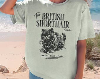 May include: A light green t-shirt featuring a black graphic of a British Shorthair cat. The text reads The British Shorthair Collective, Gentle - Calm - Plush, and La Maison Du Chat. The shirt has short sleeves and a crew neckline.