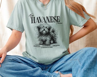 May include: A light green t-shirt featuring a black sketch of a Havanese dog. The text "The HAVANESE Collective" is above the dog, with "PLAYFUL - CHARMING - FAITHFUL" and "LA MAISON DU CHIEN" below. The shirt is made of soft material.