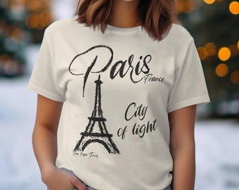 May include: A cream-colored short-sleeve t-shirt featuring a black graphic of the Eiffel Tower. The text reads Paris France, City of light, and The Eiffel Tower in a stylized script font across the front of the garment.