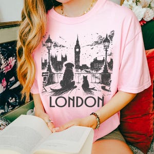 Può includere: T-shirt rosa chiaro con grafica in bianco e nero di un cane e un gatto che guardano Londra, con il Big Ben sullo sfondo. La parola "LONDON" è stampata in grandi lettere nere sotto.