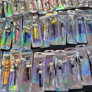 Könnte beinhalten: Eine Sammlung dekorativer Stifte in transparenten Kunststoffverpackungen. Die Stifte weisen verschiedene farbenfrohe Designs auf, darunter Perlen und Tiermuster. Jeder Stift ist mit einer kleinen Karte mit der Aufschrift "Thank you" verpackt.