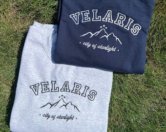 Puede incluir: Dos sudaderas, una gris y otra azul marino, con el mismo diseño. El diseño presenta el texto "VELARIS" en un contorno blanco con una cordillera blanca y tres estrellas debajo. El texto "city of starlight" está escrito en una fuente más pequeña debajo de la cordillera.