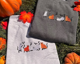 May include: Two sweatshirts with embroidered images of cats dressed as ghosts and pumpkins. The sweatshirts are gray and light gray. The sweatshirts are lying on a bed of green grass with fall leaves and pumpkins.