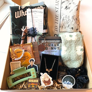 May include: A brown cardboard box filled with various items, including a book, a reading list, a hot chocolate packet, a necklace, a pair of socks, a sticker, and a hair clip. The box is decorated with a brown paper and has a brown ribbon.