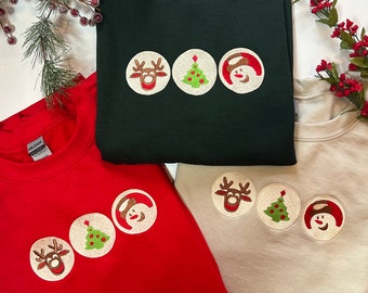 May include: Three Christmas-themed sweatshirts in green, red, and beige. Each sweatshirt features a circular design with embroidered images of a reindeer, a Christmas tree, and a snowman.
