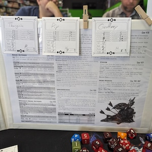 May include: A close-up of a tabletop role-playing game (RPG) session. Three character sheets are visible, with the names Thargrim, Pip, and Geoffrey. The character sheets include stats like AC, Ref, Ret, Fort, and Wee. The game board shows a detailed description of the "Cover" action, with a graphic illustration of a character hiding behind a pile of arrows. The image also includes a variety of dice, including a d20, d12, d10, d8, d6, and d4.