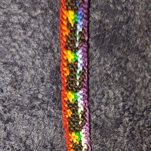 May include: A handmade friendship bracelet with a rainbow pattern. The bracelet features a woven design with black, red, yellow, green, blue, and purple threads. It has a small, clear bead at the end and a silver clasp.