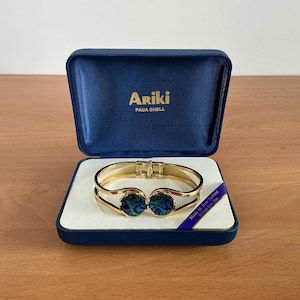 May include: A gold-toned hinged bracelet with two oval abalone shell insets, displayed in a blue velvet box. The box is open, revealing the bracelet on a white satin bed. The box is labeled "Ariki PAUA SHELL".