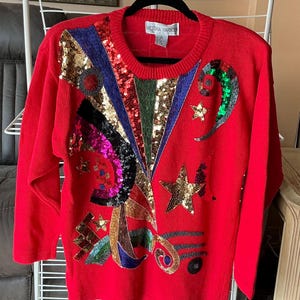 May include: A red sweater with a crew neck and long sleeves. The sweater is embellished with sequins and beads in gold, blue, green, and pink. The label displays the brand name "VICTORIA HARBOUR".