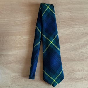 May include: A necktie featuring a dark blue and forest green plaid pattern with thin, bright yellow diagonal lines intersecting across the fabric. The tie is folded over itself, displaying a pointed tip and a smooth, woven textile construction.
