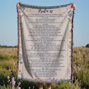 Pode incluir: Cobertor creme com o texto "Salmo 97" e um versículo bíblico impresso em azul escuro. O cobertor tem uma borda floral e uma borda com franjas. O fundo é um campo de flores silvestres.