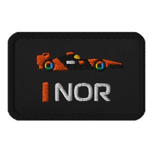 Może przedstawiać: Czarna, haftowana naszywka z pomarańczowo-czarnym, stylizowanym samochodem wyścigowym z tęczowymi kołami. Tekst "INOR" jest wyhaftowany na biało i pomarańczowo. Naszywka ma czarną obwódkę.