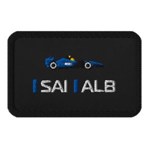 Op de afbeelding: Geborduurde patch met een zwarte achtergrond. Een blauwe raceauto is afgebeeld aan de bovenkant, met de tekst "I SAI ALB" eronder. De tekst en de auto zijn respectievelijk in wit en blauw. De patch heeft een zwarte rand.