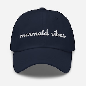 Op de afbeelding: Een marineblauwe baseballpet met witte geborduurde tekst die "mermaid vibes" luidt met twee kleine hartjes boven het woord "vibes".