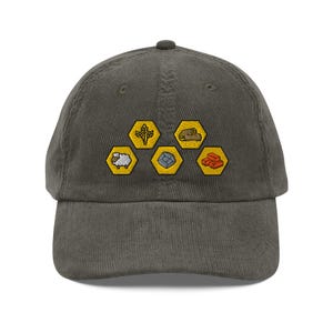 Puede incluir: Gorra de béisbol de pana verde oliva. Cuenta con parches hexagonales amarillos bordados con trigo, troncos, ladrillos, una oveja y una piedra. La gorra tiene una visera curva y una correa ajustable.