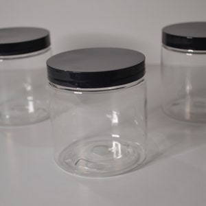 Op de afbeelding: Drie doorzichtige plastic potten met zwarte deksels. De potten zijn leeg en hebben een gladde, afgeronde vorm.