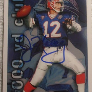 May include: A trading card featuring a football player in a blue and white uniform with the number 12. The player is throwing a brown football. The card has the text "3000 yd club".