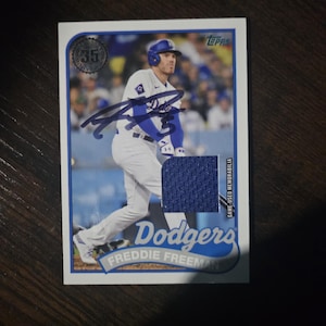 May include: A Topps baseball card featuring Freddie Freeman of the Dodgers in a white and blue uniform. The card includes a blue ink autograph, a blue fabric jersey swatch, and text reading Dodgers, Freddie Freeman, and Game-Used Memorabilia.