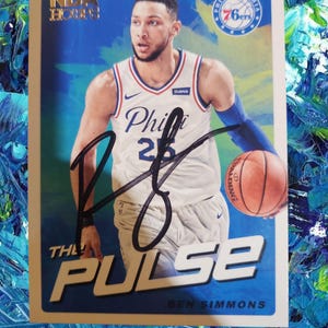Puede incluir: Una tarjeta de baloncesto con Ben Simmons con el uniforme de los 76ers de Filadelfia, sosteniendo un balón de baloncesto. La tarjeta tiene un fondo azul con el texto "THE PULSE" y "NBA Hoops". La tarjeta está autografiada.