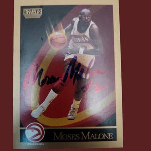 Peut inclure: Une carte de collection de basket-ball SkyBox représentant Moses Malone portant un uniforme rouge et or des Atlanta Hawks. La carte a un fond doré et une bordure dorée. La carte est signée en encre bleue par Moses Malone.