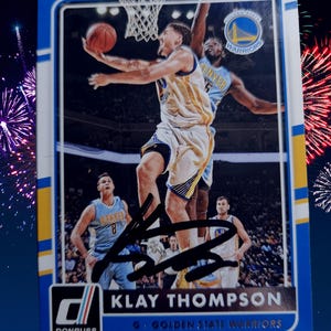 May include: A Klay Thompson basketball card from Donruss showing the Golden State Warriors player in action. The card has a blue border and an autograph. The background features other players in a game.