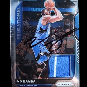May include: A basketball trading card featuring Mo Bamba of the Orlando Magic in a shooting pose. The card has a blue and silver design with the word "Sensational" and a patch of blue fabric. The card is signed.