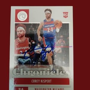 Peut inclure: Une carte de collection de basketball représentant Corey Kispert, un joueur recrue des Washington Wizards. La carte est de la série Panini Chronicles et présente une photo de Kispert en uniforme des Wizards, dribblant le ballon. La carte a un fond argenté avec des accents rouges et le texte "Chronicles" en grandes lettres grasses. La carte comprend également le nom, le numéro et l'équipe de Kispert.