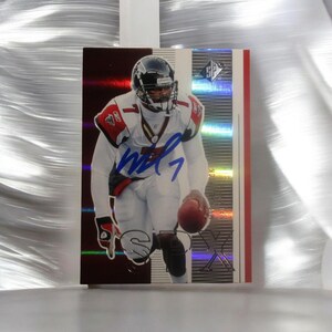 May include: A rectangular sports trading card featuring a football player in a white and red uniform with a black helmet, holding a brown football. The card has a metallic red and silver background with a blue ink autograph and the SP logo at the top.