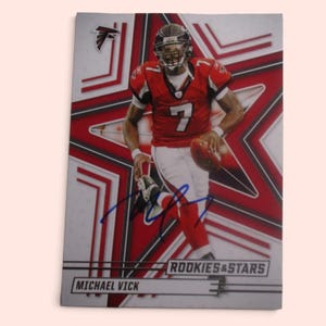 May include: A rectangular sports trading card featuring Michael Vick in a red and white Atlanta Falcons uniform with the number 7. The card has a blue ink autograph, a red star background, and the text Rookies & Stars and Michael Vick.