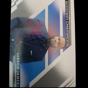 Peut inclure: Carte de collection de basket-ball avec LeBron James en costume bleu marine. La carte présente un motif géométrique bleu et blanc avec le texte "GOODWIN CHAMPIONS" et "LEBRON JAMES BASKETBALL". La carte comporte également une signature rose.