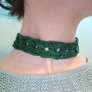 May include: A dark green crochet choker necklace. The necklace is made of a woven pattern and sits around the neck. The choker is a solid, deep green color and is worn over a gray sweater.
