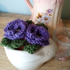 Könnte beinhalten: Zwei lila gehäkelte Blumen mit grünen gehäkelten Blättern in einer weißen, vogelartigen Keramik-Schale.