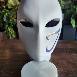 May include: White mask with a purple and gold design. The mask is sitting on a white circular stand.
