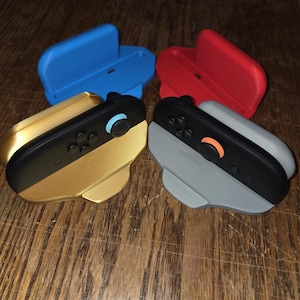 May include: Four game controller accessories in the image. Two controllers are black and gold, and black and gray. Two stands are blue and red. The controllers have buttons and joysticks.