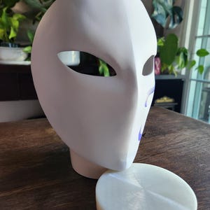 May include: A white plaster mask with two eye holes and a smooth surface. The mask is sitting on a white round base.