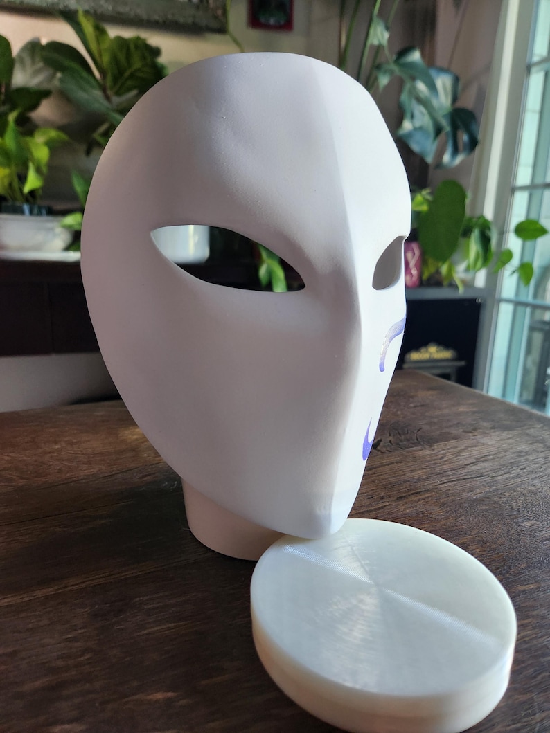 May include: A white plaster mask with two eye holes and a smooth surface. The mask is sitting on a white round base.