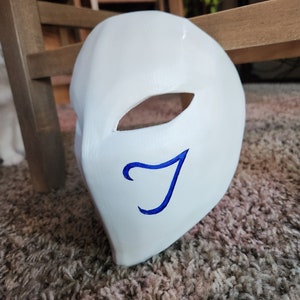May include: A white mask with a blue letter 'J' painted on it. The mask has a single eye hole.